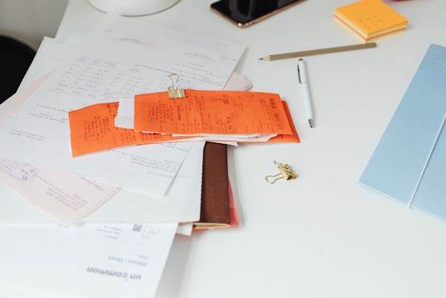 Vista en ángulo superior de un escritorio blanco con diversos documentos administrativos esparcidos. En el centro destaca un fajo de tickets o recibos de color naranja sujetos con una pinza metálica, apilados sobre folios impresos. Alrededor hay un bolígrafo blanco, un lápiz, un teléfono móvil, notas adhesivas amarillas y una carpeta azul claro. Este papeleo contable ilustra la gestión documental y los gastos que suelen analizarse al explicar la diferencia entre domicilio social y fiscal de un negocio.