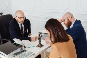 Un abogado calvo con gafas y traje oscuro hablando seriamente desde su escritorio blanco con dos clientes, una mujer con chaqueta color camello y un hombre con barba, durante una reunión de asesoramiento legal en un domicilio social, con una estatua de la justicia y un libro abierto sobre la mesa.