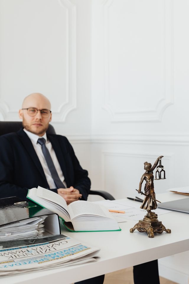 Primer plano nítido de una pequeña estatua de bronce de la dama de la justicia, un libro grueso abierto y carpetas con documentos sobre un escritorio blanco, mientras un abogado calvo con gafas y traje oscuro posa desenfocado al fondo en la oficina de un domicilio social.