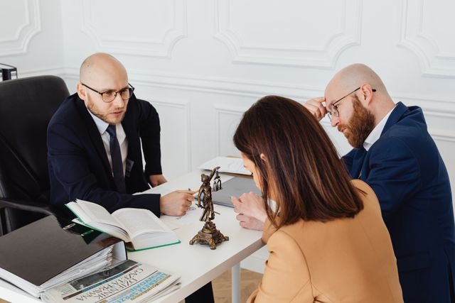 Un abogado calvo con gafas y traje oscuro hablando seriamente desde su escritorio blanco con dos clientes, una mujer con chaqueta color camello y un hombre con barba, durante una reunión de asesoramiento legal en un domicilio social, con una estatua de la justicia y un libro abierto sobre la mesa.