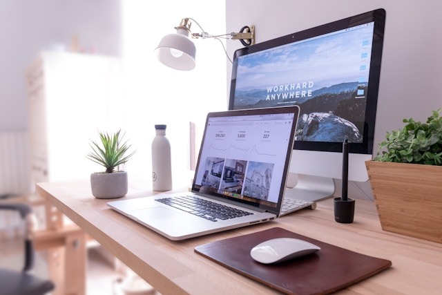 Vista de un escritorio de madera clara muy iluminado. Al fondo hay un monitor de escritorio que muestra un paisaje montañoso con la frase "WORKHARD ANYWHERE", y delante una laptop abierta exhibiendo una página web con gráficos y estadísticas. También se observa un ratón blanco sobre una alfombrilla de cuero marrón, una pequeña maceta con una planta, una botella blanca y una lámpara de pinza, ilustrando un entorno de trabajo digital ideal al domiciliar sociedad.