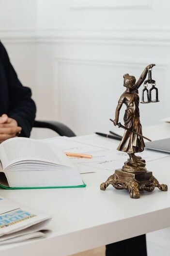 Primer plano nítido de una pequeña estatua de bronce de la dama de la justicia, un libro grueso abierto y carpetas con documentos sobre un escritorio blanco, mientras un abogado calvo con gafas y traje oscuro posa desenfocado al fondo en la oficina de un domicilio social.